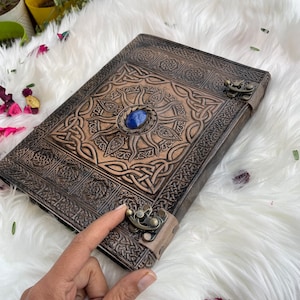 May include: A brown leather journal with a Celtic knot design and a blue gemstone in the centre. The journal has a clasp closure and a strap.