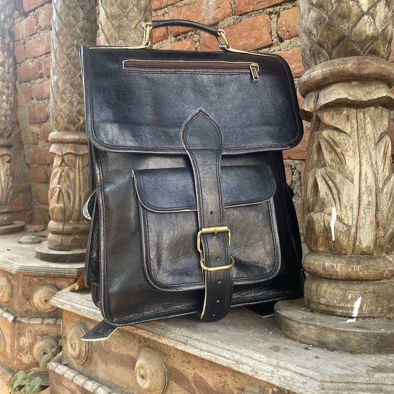 May include: A black leather backpack with a large front pocket and a buckle closure. The backpack has a top handle and adjustable straps.
