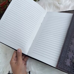 May include: A brown leather journal with a detailed embossed design on the cover. The journal is open to reveal lined pages.