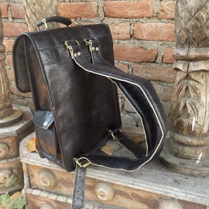 May include: A black leather backpack with a flap closure and a front pocket. The backpack has a white trim around the edges of the straps and a brass buckle.