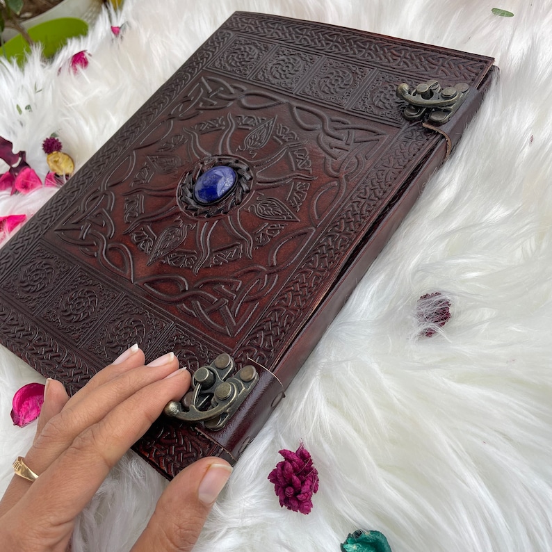 May include: A brown leather journal with a Celtic knot design and a blue gemstone in the centre. The journal has a metal clasp and is closed.