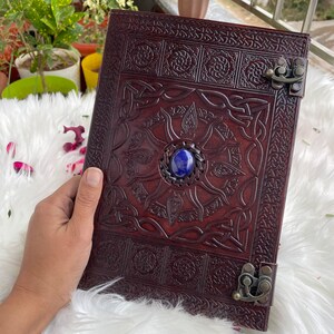 May include: A brown leather journal with intricate Celtic knotwork designs and a blue gemstone embellishment. The journal has two metal clasps.