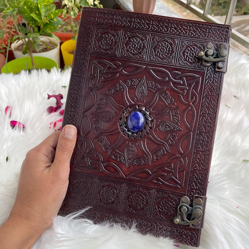 May include: A brown leather journal with intricate Celtic knotwork designs and a blue gemstone embellishment. The journal has two metal clasps.