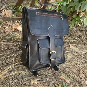 May include: A black leather backpack with a large front pocket and a buckle closure. The backpack has a top handle and adjustable straps.
