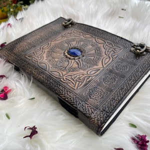 May include: A brown leather journal with a Celtic knot design and a blue gemstone in the centre. The journal has metal clasps and is decorated with intricate details.