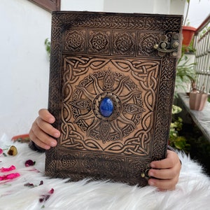 May include: A brown leather journal with intricate Celtic knotwork designs and a blue gemstone embedded in the centre. The journal has a metal clasp and is closed.