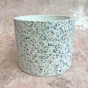 May include: A cylindrical planter with a speckled terrazzo design. The planter features a light cream base with embedded chips of grey, black, and reddish-brown. The interior is a solid white colour. Suitable for indoor or outdoor use.