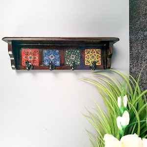 May include: A dark brown wooden wall shelf with four decorative ceramic tiles and black metal hooks. The tiles feature colorful floral patterns in red, blue, green, and yellow. The shelf is mounted on a light gray wall.