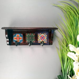 May include: A dark brown wooden wall shelf with three decorative ceramic tiles and three black metal hooks. The tiles feature colorful floral patterns in orange, blue, green, and yellow. The shelf is mounted on a gray wall.
