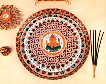 May include: A circular decorative tray featuring a central illustration of the deity Ganesha, surrounded by intricate red, green, and gold patterns. The set includes a matching floral-shaped coaster, a small bell, and five black incense sticks.