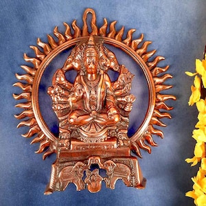 May include: A copper-colored Hindu deity statue of Hanuman, the monkey god, with four arms, sitting in a meditative pose. The statue is surrounded by a circular sunburst design and sits on a base with two elephants.