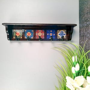 May include: A dark brown wooden shelf with five black hooks. The shelf features five decorative tiles with floral patterns in red, blue, and yellow. The shelf is mounted on a gray wall.
