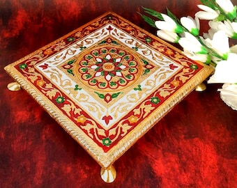May include: A square decorative platform featuring a central circular floral motif in red, green and gold. The surface is adorned with intricate white and gold filigree patterns and a red border. It stands on four small, rounded gold-toned feet.