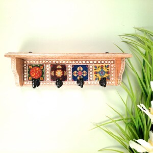 May include: A wooden shelf with four black hooks, featuring colorful square tiles with floral patterns. The shelf is painted with a light brown color and has a decorative border. The tiles have red, brown, blue, and yellow designs.