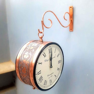 May include: A copper-colored clock with a white face and black Roman numeral markings. The clock is hanging from a copper-colored hook and has a decorative floral pattern on the side. The clock face reads "Village Clocks Birmingham est 1882".