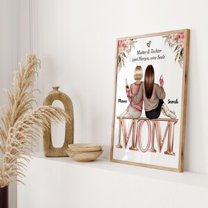 May include: A framed illustration of two women sitting on the word "MOM" in rose gold letters. The women are holding glasses of wine and the text above them reads "Mutter & Tochter zwei Herzen, eine Seele". The names "Mara" and "Sarah" are written below the women.