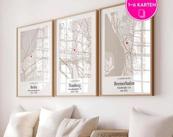 May include: Three framed city map prints with a neutral color palette. Each map features a red heart marking a location, with city names and dates below. The prints are displayed above white pillows and a woven basket.