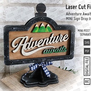 以下が含まれることがあります： 黒枠で古びた木製の看板。看板には「Adventure awaits」という文字と、その裏には山々の風景が描かれている。看板は黒色の台座に置かれ、青と白の市松模様のリボンが結ばれている。