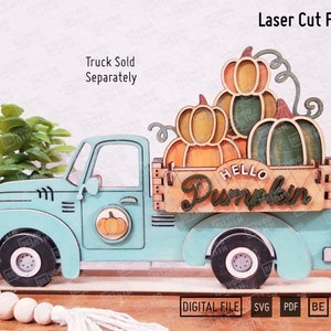 May include: A laser-cut wooden teal truck carrying pumpkins. The truck bed features a sign that reads "Hello Pumpkin." The pumpkins are in shades of orange and green. The text "Truck Sold Separately" is above the truck.