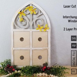 Op de afbeelding: Een wit houten raamkozijn met een decoratief ontwerp, met vier vierkante vakken. Het frame is versierd met gele bloemen. De tekst "Laser Cut Files", "Interchangeable Window", "2 Layer Project", "SVG", "PDF", "DIGITAL FILE", "BE", "YOO", en "NIK" is zichtbaar aan de rechterkant van de afbeelding.