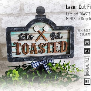 May include: A black and white sign with the text "Let's get TOASTED" in brown and white letters. The sign has a rustic woodgrain design and features a pair of crossed marshmallows with a campfire behind them. The sign is on a black stand with a blue and white checkered bow.