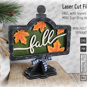 May include: A wooden sign with the word "fall" in cursive, featuring orange leaf accents. The sign is framed in a dark gray color and sits on a wooden base. The sign includes the text "Laser Cut Files" and "MINI Sign Drop In".
