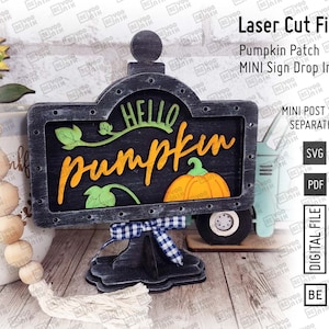 Op de afbeelding: Een zwart houten bord met een wit en blauw ruitjeslint, met de tekst "Hello Pumpkin". Het bord staat op een standaard en heeft een pompoen-uitsparing met oranje en groene details. Het bord is onderdeel van een laser-snijbestandset voor een mini-bord voor een pompoenveld.