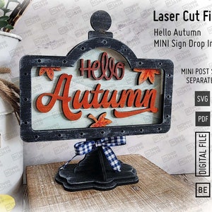 May include: A laser cut wooden sign with the words "Hello Autumn" in a decorative font. The sign is on a black stand with a white and blue checkered ribbon. The sign is designed to be used as a drop-in for a larger sign.
