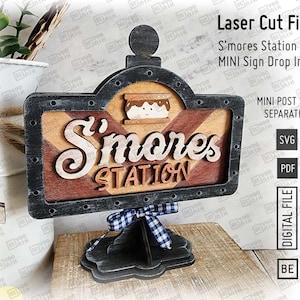 May include: A black and brown wooden sign with the text "S'mores Station" in white lettering. The sign is attached to a black stand with a blue and white checkered ribbon. The sign is a laser cut file for crafting.