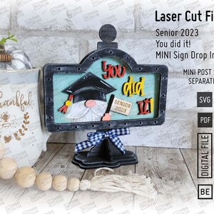 May include: A laser cut wooden sign with a graduation cap and the text "You did it!" and "Senior 2023". The sign is on a black stand with a blue and white checkered ribbon.