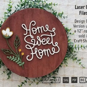 May include: A wooden round sign with the words "Home Sweet Home" in white script. The sign is decorated with a floral design featuring white flowers and green leaves.