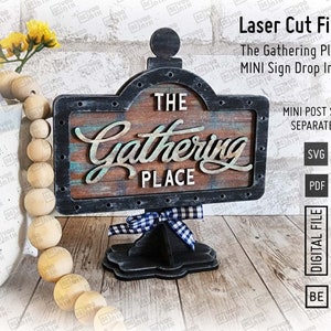 Puede incluir: Diseño de archivo de corte por láser para un letrero de madera que dice "The Gathering Place". El letrero es de estilo rústico con un acabado negro y marrón. El letrero está en un soporte negro con una cinta a cuadros blancos y negros.