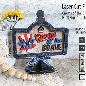 May include: A laser cut wooden sign with a gnome wearing a red, white, and blue hat. The sign says "Gnome of the Brave" in red, white, and blue letters. The sign is on a black stand with a blue and white checkered bow.