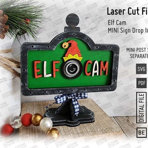 Può includere: Un file di taglio laser per un cartello natalizio che dice "Elf Cam" con uno sfondo verde e una cornice nera. Il cartello è su un supporto con un nastro a scacchi rosso e bianco. Il file è disponibile nei formati SVG e PDF.