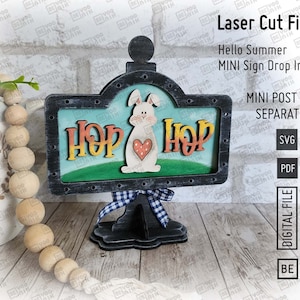 May include: A laser cut wooden sign with a bunny rabbit and the words "Hop Hop" in a whimsical font. The sign is painted in shades of green, blue, and white and has a black base. The sign is designed for a mini post, which is sold separately.