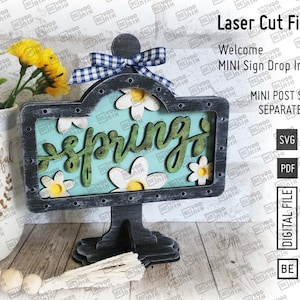 May include: A laser cut wooden sign with a blue and white checkered bow, a blue background, and the word "spring" in white letters. The sign is decorated with white flowers and has a black stand.