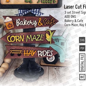 May include: Three wooden signs with arrows pointing to the right. The signs are painted in a rustic style with brown, orange, and yellow paint. The signs read "Bakery & Cafe", "Corn Maze", and "Hay Rides".
