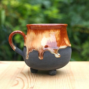 Könnte beinhalten: Eine handgefertigte Keramiktasse mit einem einzigartigen Design. Die Tasse hat einen dunkelgrauen Boden mit drei kleinen Füßen, der in eine hellbraune und orangefarbene glasierte Oberseite übergeht. Die Glasur tropft an den Seiten herunter und erzeugt einen strukturierten Effekt. Der Griff ist ebenfalls dunkelgrau.
