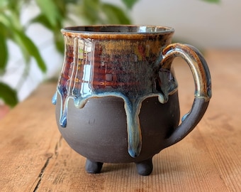May include: A ceramic mug with a rounded body and three small feet. The upper portion features a glossy, multi-toned glaze in shades of amber, brown, and blue that drips down over the matte, dark brown base. It includes a curved handle.