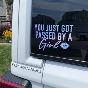 May include: A white vehicle's rear window displays a holographic sticker. The sticker reads "YOU JUST GOT PASSED BY A Girl" in white and blue-purple gradient lettering, with a lip graphic. Below, the text "ETSY: 4X4ADVENTURES" is printed in black.