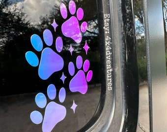 May include: A car window with a sticker of four paw prints in a gradient of blue and purple. The paw prints are arranged in a cluster with a few small stars. The text "Etsy: 4x4adventures" is visible on the window.