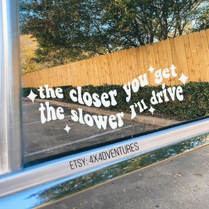 May include: White vinyl sticker with the text "the closer you get the slower I'll drive" in a retro font with sparkling stars on a car window. The text is reflected in the window. The sticker is from Etsy shop 4x4adventures.
