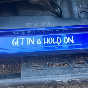 May include: A blue car door with a white vinyl decal that reads "GET IN & HOLD ON". The decal is on the lower part of the door, above a black step.