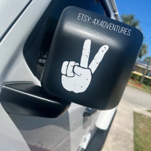 May include: A black side view mirror with a white peace sign sticker. The sticker has a distressed look and the text "ETSY: 4X4ADVENTURES" is visible on the mirror.