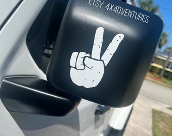 May include: A black side view mirror with a white peace sign sticker. The sticker has a distressed look and the text "ETSY: 4X4ADVENTURES" is visible on the mirror.