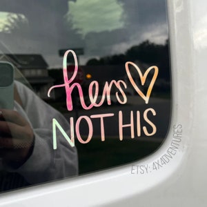 May include: A pink and orange iridescent vinyl sticker with the text "hers not his" and a heart. The sticker is on a car window.