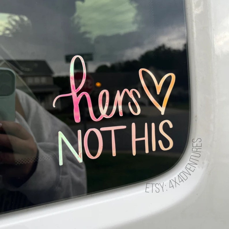 May include: A pink and orange iridescent vinyl sticker with the text "hers not his" and a heart. The sticker is on a car window.