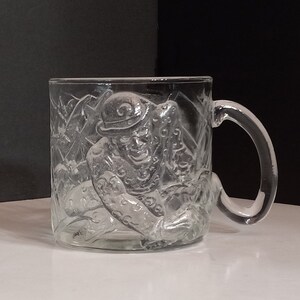 May include: A clear glass mug with a raised design of the Joker from Batman. The mug has a handle and is perfect for enjoying a hot beverage.