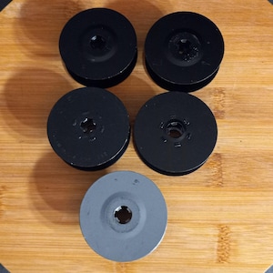 May include: Five black and gray film reels are arranged on a wooden surface. Four reels are black, and one is gray. The reels have a circular design with a central hole.