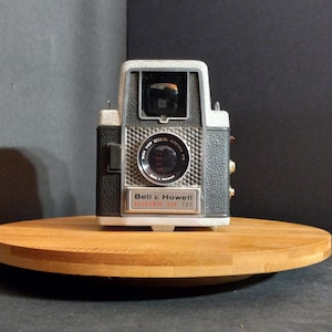 May include: A vintage Bell & Howell Electric Eye 127 camera featuring a grey and black textured body, a central lens with a metallic grid surround, and a top-mounted viewfinder. The front plate displays the brand name and model text clearly.